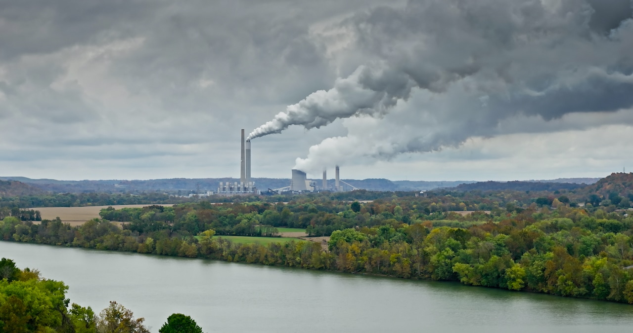 Drone shot looking across the Ohio River and the farmland and forest landscape on the West Virginia side towards Cheshire, Ohio. In the distance, Kyger Creek Power Plant and Gavin Power Plant are sending up plumes of smoke and steam into the overcast sky.